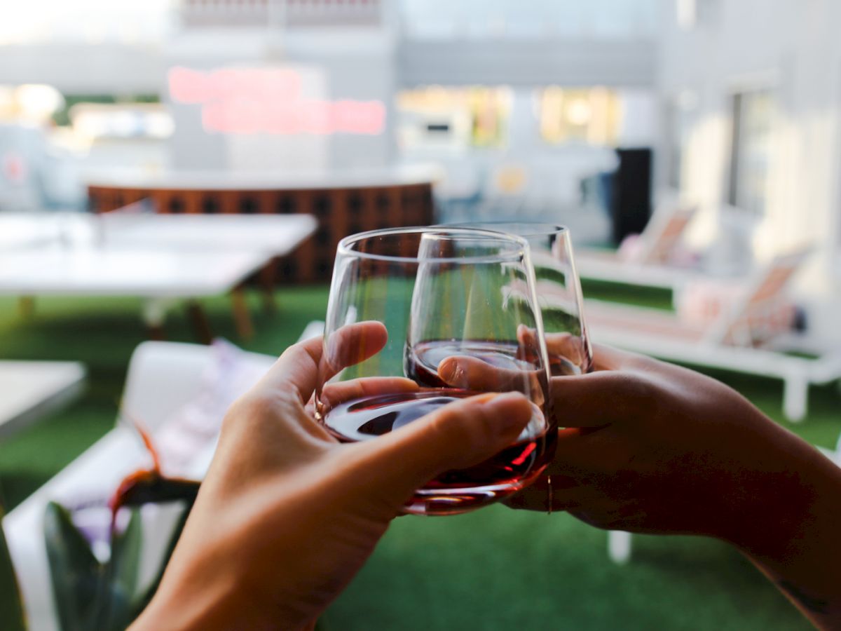 Two people clinking glasses with a blurred outdoor lounge area in the background, featuring greenery and white furniture.