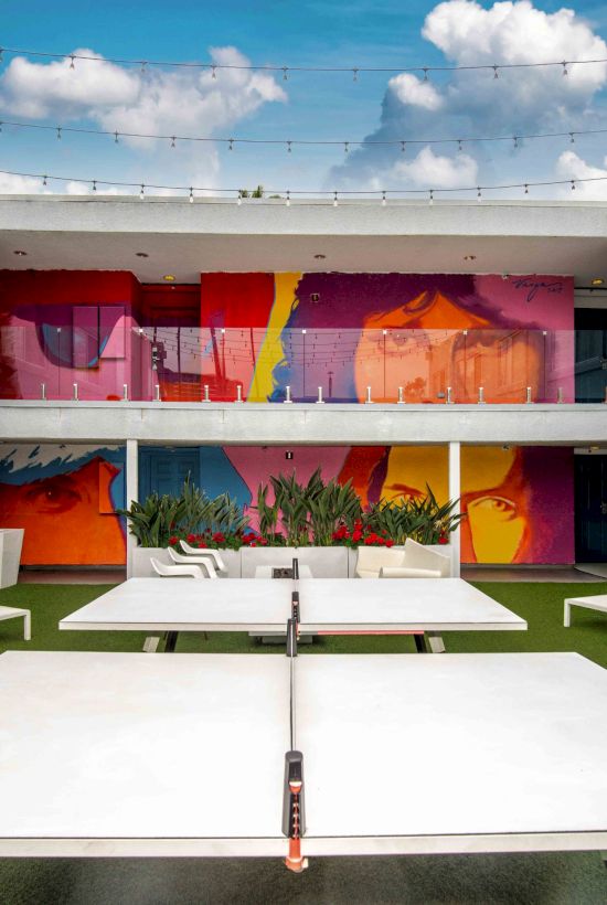 The image shows a courtyard with lounge chairs, a ping pong table, colorful mural walls, and decorative cushions under a blue sky.