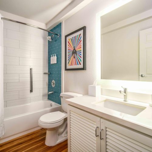 A modern bathroom with a shower, toilet, sink, and mirror. There's a colorful picture on the wall and a towel on the counter.
