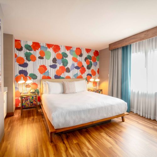 A modern hotel room with a colorful accent wall, bed, bedside lamps, and a large window with curtains, creating a cozy atmosphere.
