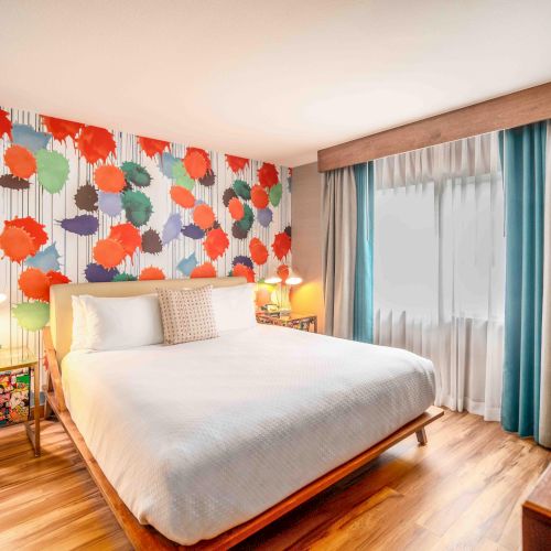 A colorful bedroom with a double bed, vibrant polka-dotted wall, bedside tables, and curtains covering a window.