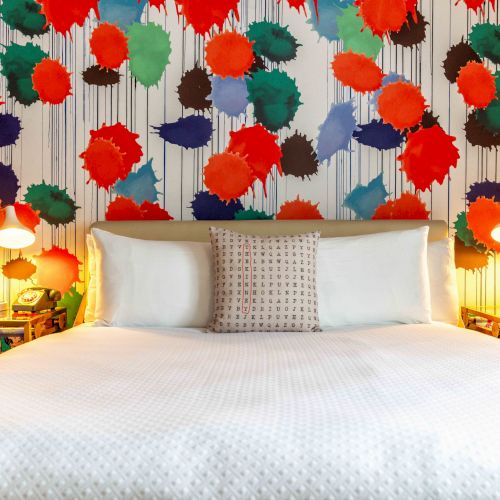 A bed with white bedding is set against a colorful, abstract floral wall. Flanked by two bedside tables with lamps, adding a warm ambiance.