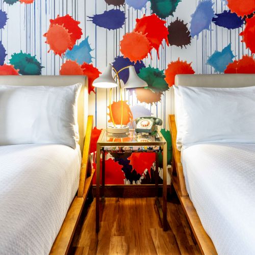 A colorful bedroom with two beds, a vibrant splatter-paint wallpaper, and a small nightstand with a lamp and retro phone between them.