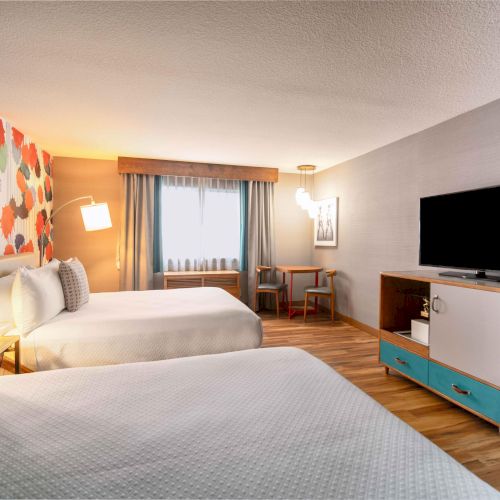 A hotel room with two beds, a colorful wall, a TV on a stand, a table with chairs by the window, and wooden flooring.