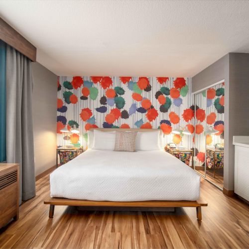 A colorful hotel room with a bed, vibrant floral wallpaper, wooden floor, white cabinets, and a window with teal curtains.