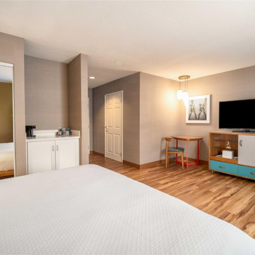 A modern hotel room with a bed, TV, a small table with a chair, and a mirrored wardrobe. Warm lighting and wood floors complete the scene.