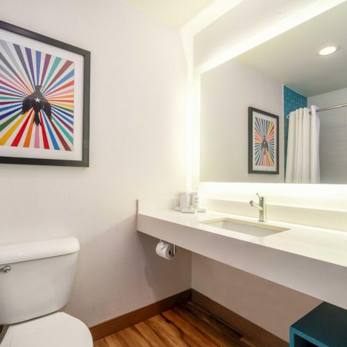 A modern bathroom with a toilet, a large mirror above a sink, towels, and colorful framed art on the wall, featuring a radial design.