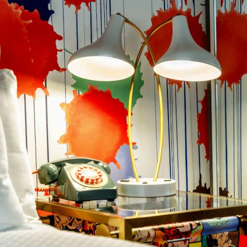 Colorful wallpaper with splotches, vintage rotary phone, double lamps, and a bed edge on a bedside table.