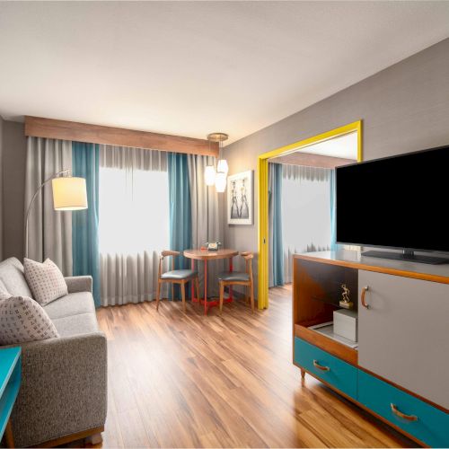 The image shows a modern living room with a sofa, TV, table, and chairs. Decor includes bright accents and a window with curtains.