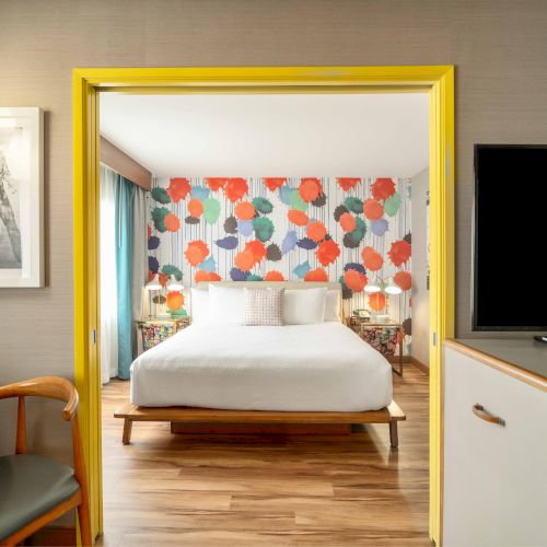 The image shows a modern bedroom with colorful wallpaper, a white bed, artwork, and a TV on the right.