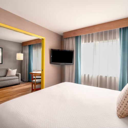 A modern hotel room with a bed, wall-mounted TV, window with curtains, and a separate sitting area with a sofa and table.