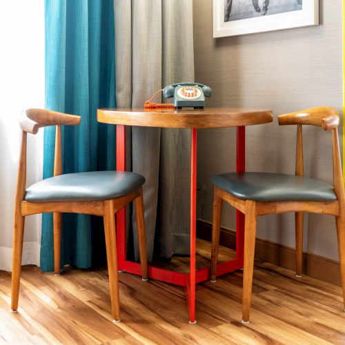 A small round table with two chairs, an old-fashioned rotary phone on top, and colorful curtains in the background by the window.