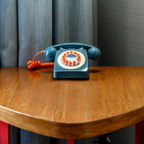 A retro rotary dial telephone with an orange cord is placed on a wooden triangular table near a curtain and wall.