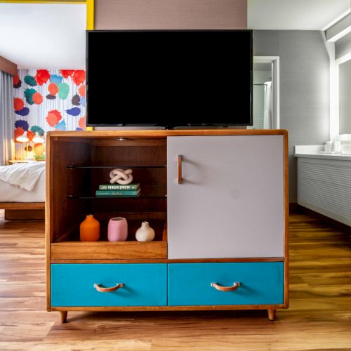 The image shows a hotel room with a colorful cabinet, TV, bed, and decorative wall art, featuring a mix of wood and bright colors.