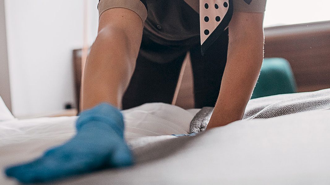 A person is making a bed, wearing a blue glove and a uniform, smoothing out the sheets in a room.