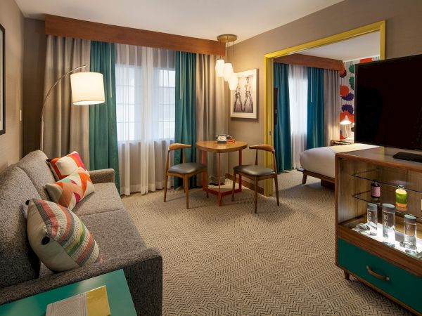 The image shows a modern hotel room with a sofa, table, chairs, TV, and bed. Bright accents and contemporary decor complete the space.