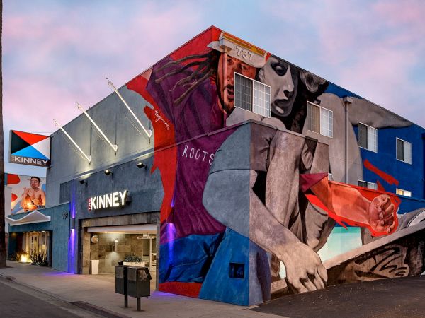 A building with vibrant mural art depicting people and abstract shapes, under a pink sky, labeled "KINNEY."