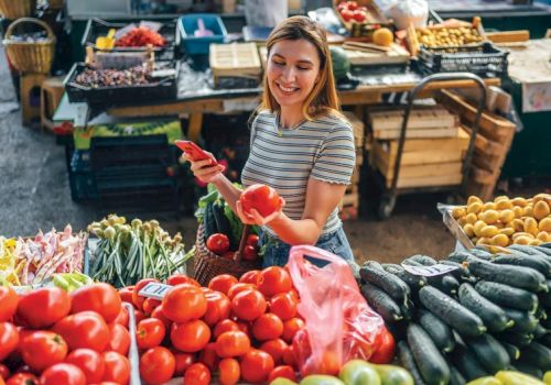 A person is choosing tomatoes at a market stall with various fresh vegetables, smiling and enjoying the shopping experience.