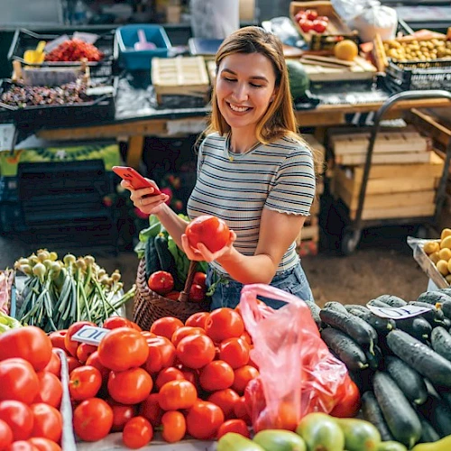 A person is choosing tomatoes at a market stall with various fresh vegetables, smiling and enjoying the shopping experience.