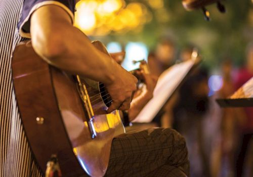 A person is playing an acoustic guitar, surrounded by warm, blurred lights in the background, creating a cozy atmosphere.