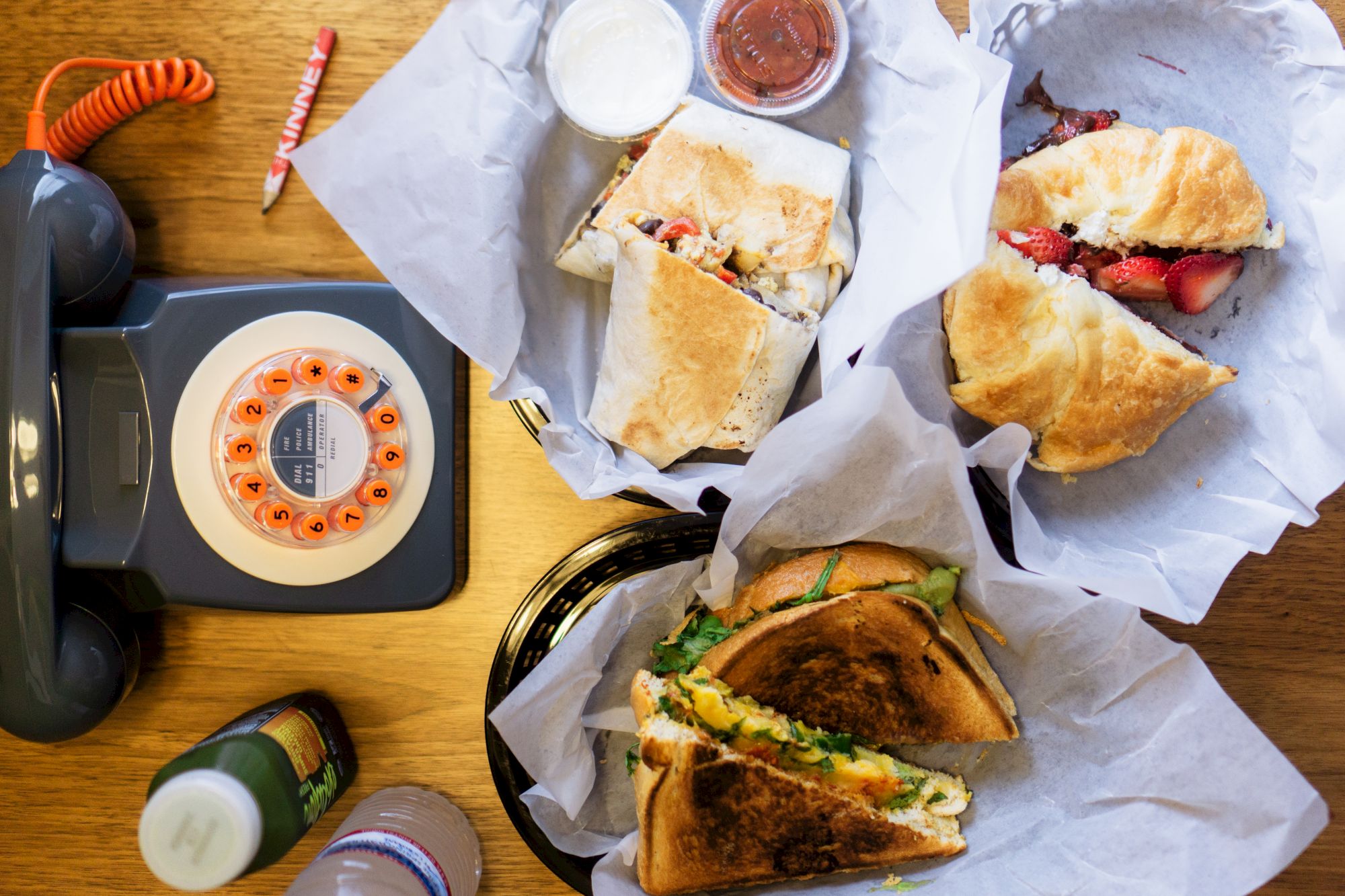 An overhead view of food, including wraps and sandwiches, with a retro rotary phone and drinks on a wooden table.