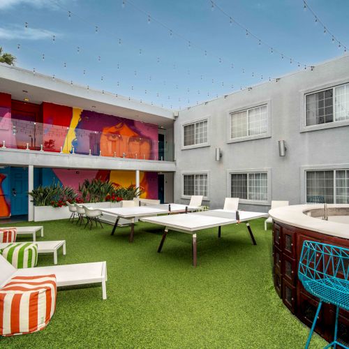 A modern courtyard with lounge chairs, a ping pong table, colorful wall art, string lights above, and a seating area with vibrant decor.