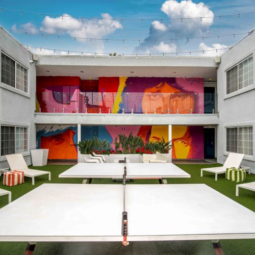 A modern courtyard with white lounge chairs, a white ping pong table, colorful mural, and string lights overhead under a blue sky.