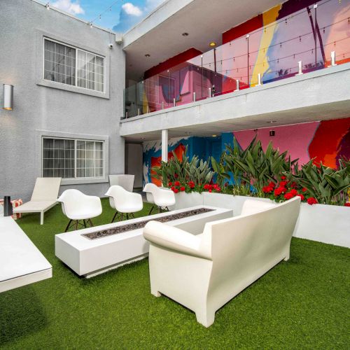 The image shows a modern outdoor patio with white seating, vibrant wall art, and greenery on an artificial grass surface.