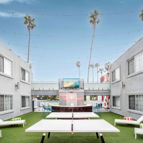 A courtyard with modern design features a ping pong table, lounge chairs, and palm trees, surrounded by a two-story building.
