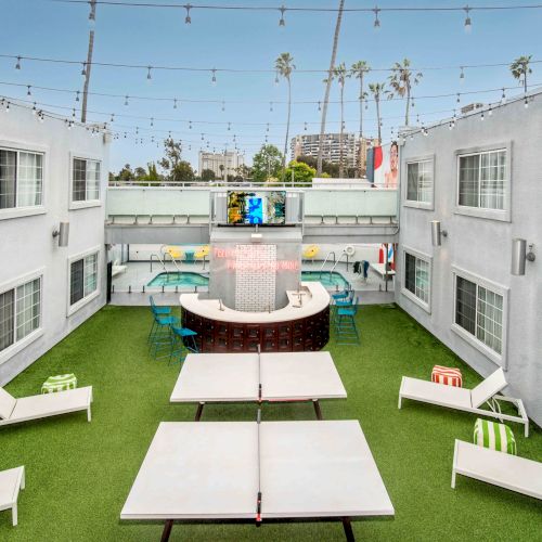 This image shows a courtyard with loungers, a ping-pong table, string lights, and palm trees, in a setting surrounded by white buildings.