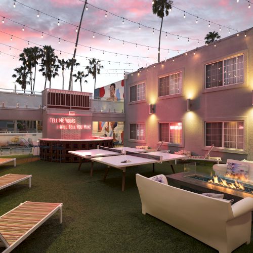 A cozy courtyard with ping-pong tables, lounge chairs, a sofa, string lights, and a fire pit, set against a pink sunset sky and palm trees.