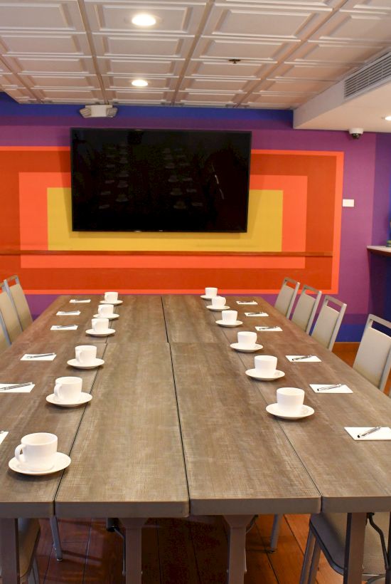 A modern meeting room with a long table, chairs, cups, saucers, a large screen on a colorful wall, and patterned wallpaper.
