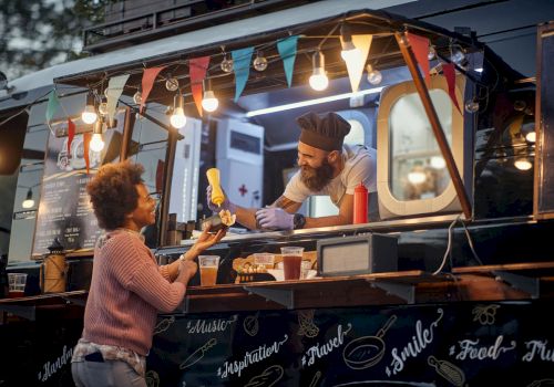 A food truck scene with a vendor in a chef's hat handing food to a customer, featuring colorful lights and decorative banners.