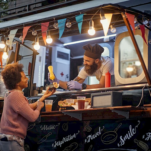 A food truck scene with a vendor in a chef's hat handing food to a customer, featuring colorful lights and decorative banners.