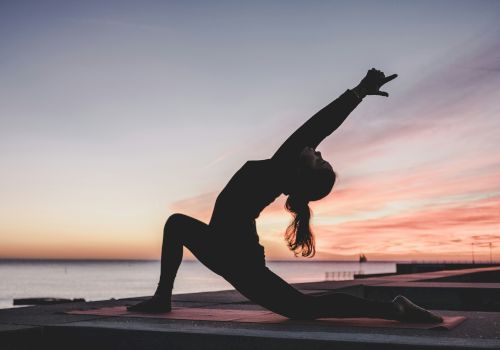 A person doing yoga in a lunge position at sunrise or sunset near the water, silhouetted against the colorful sky.
