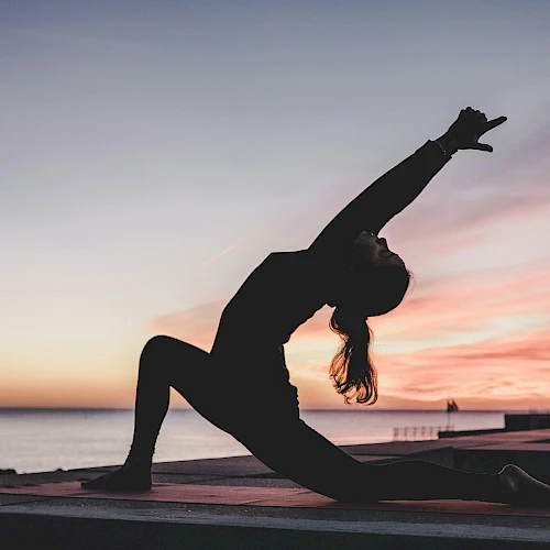 A person doing yoga in a lunge position at sunrise or sunset near the water, silhouetted against the colorful sky.