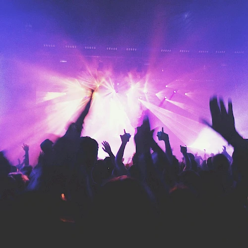 A crowd with raised hands at a concert or music event, illuminated by colorful stage lights and beams, creating a lively atmosphere.