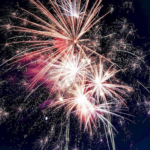 Bright fireworks burst against a dark sky, creating a dazzling, colorful display of light and sparks cascading outward.