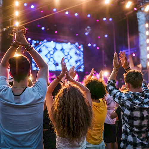 People are enjoying a live concert, clapping and cheering, with bright stage lights and a colorful backdrop in the background.