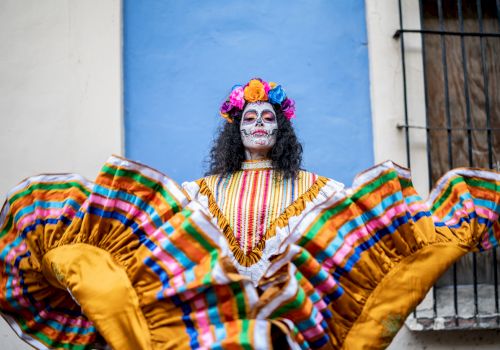 A person in colorful traditional attire and skull face paint, likely for Día de los Muertos, stands against a blue wall, holding up their skirt.