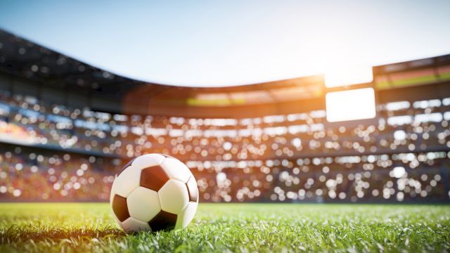 A soccer ball on a grassy field in a sunlit stadium with blurred audience in the background.