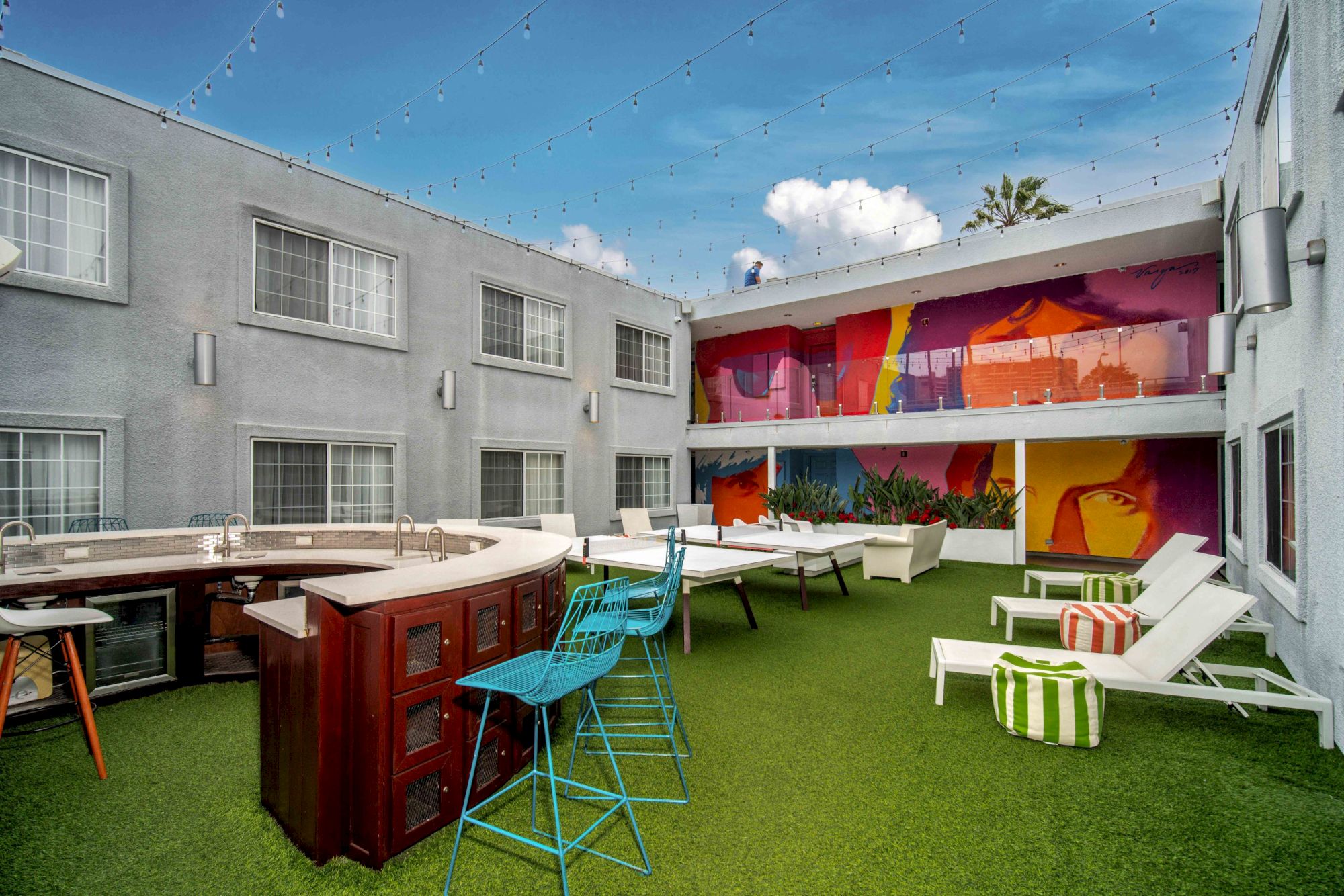 An outdoor courtyard with seating, colorful mural walls, and decorative lighting, surrounded by gray buildings under a blue sky.