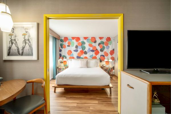 The image shows a modern hotel room with a colorful floral accent wall, a bed, and a view of a sitting area and TV.