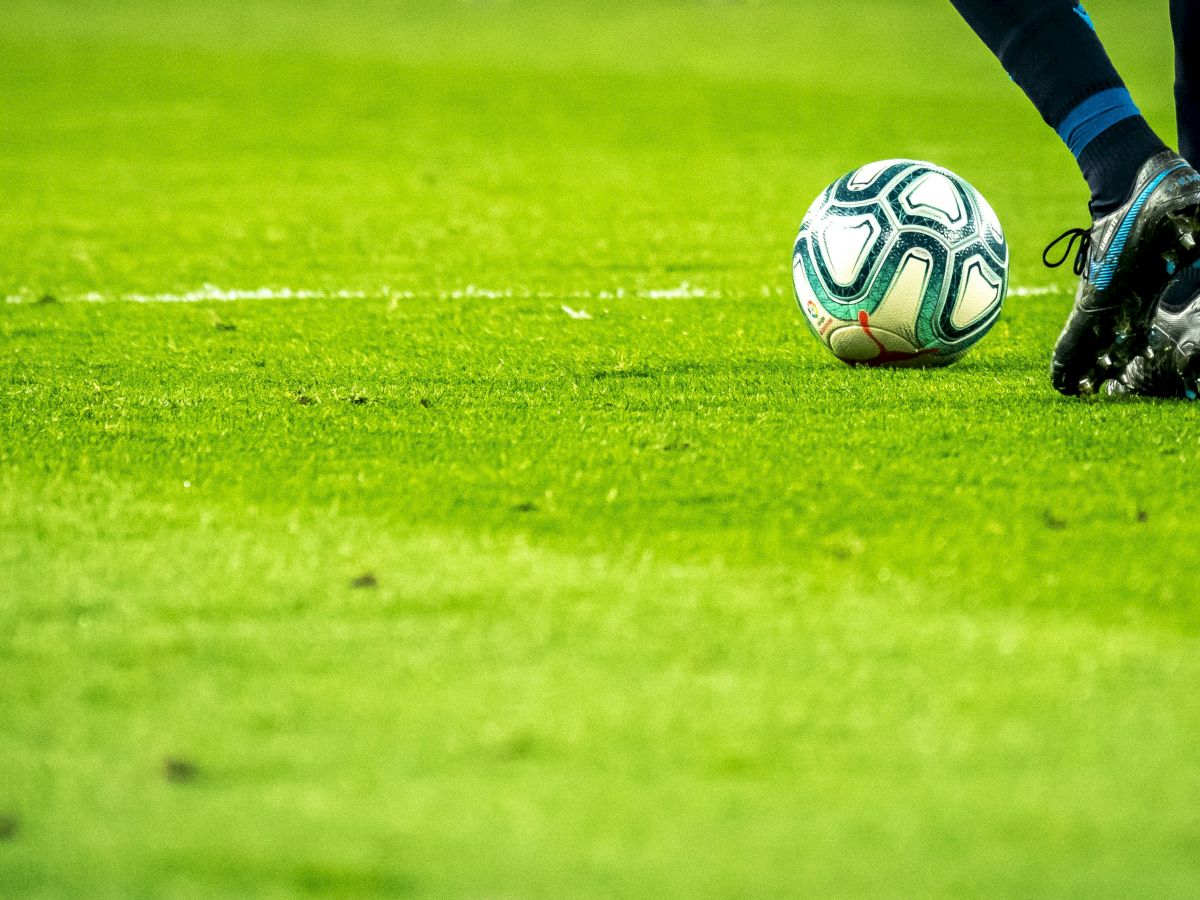 A close-up of a soccer ball and a player&rsquo;s cleats on a lush green pitch, ready for kickoff.