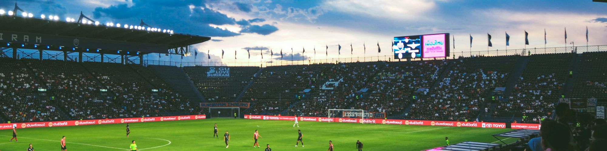 A soccer stadium filled with fans, players on the green field, dramatic evening sky, and bright stadium lights glowing as a match unfolds.