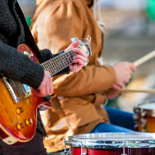 A guitarist and a drummer playing outdoors, with a focus on the guitar in the foreground and drums in the background.