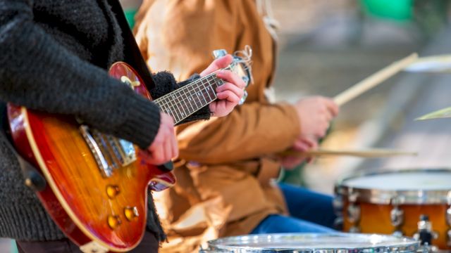 A guitarist and a drummer playing outdoors, with a focus on the guitar in the foreground and drums in the background.