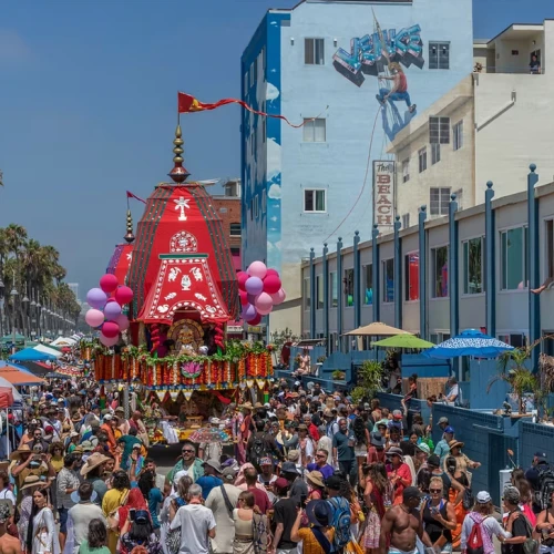 A crowded street festival with a colorful float parade, balloons, food stalls, palm trees, and a lively seaside vibe.