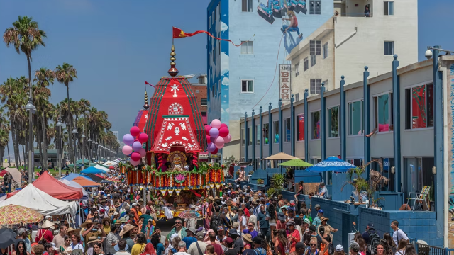 A crowded street festival with a colorful float parade, balloons, food stalls, palm trees, and a lively seaside vibe.