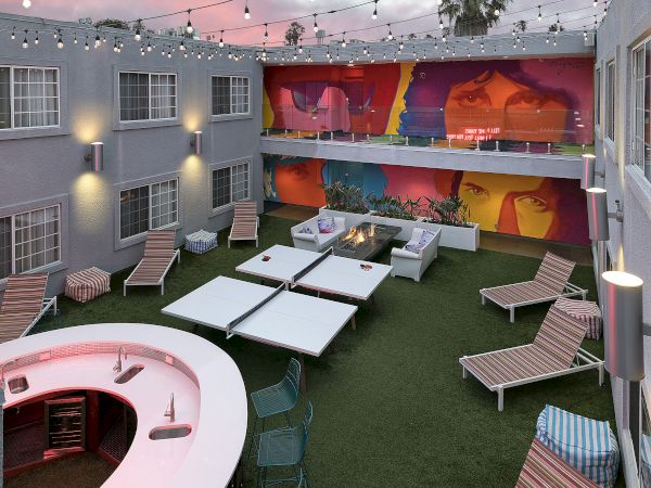 A rooftop courtyard lounge at dusk with string lights, a fire pit, striped chairs, ping-pong tables, and vibrant mural walls.
